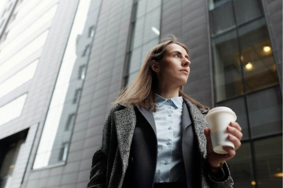Zdjęcie przedstawia businesswoman idącą z kawą na tle wieżowca