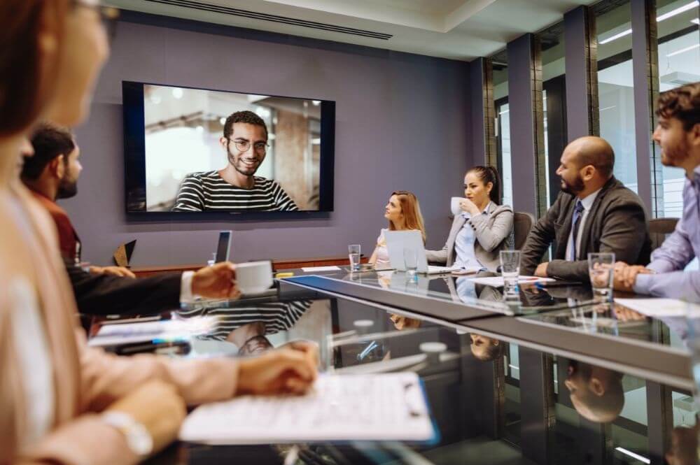 zdjęcie przedstawia konferencję w biurze, gdzie jedna osoba wdzwania się innego miejsca i jest widoczna na monitorze